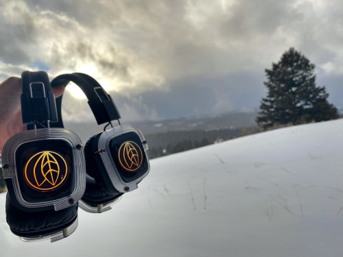 A hand holds a pair of headphones against a snowy landscape and cloudy sky, with a tree in the background.
