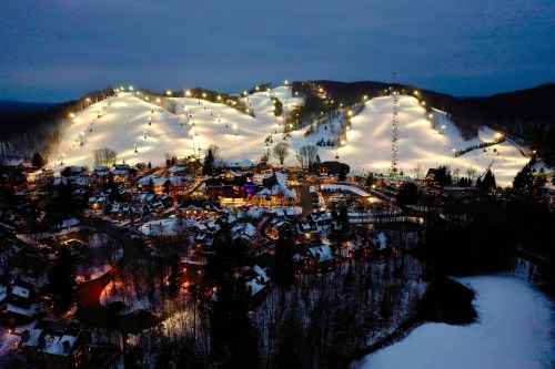 A snowy landscape at dusk, featuring a ski resort with illuminated slopes and a nearby village.