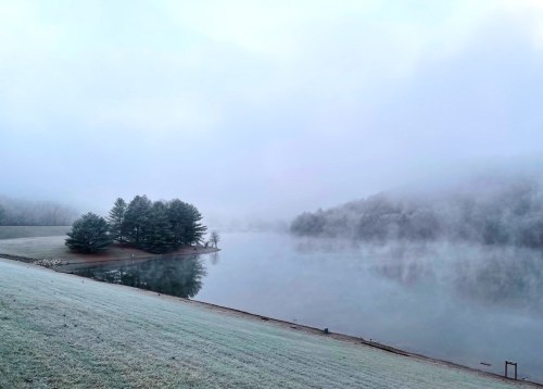 A serene, foggy landscape featuring a calm lake, frosted grass, and an island with trees in the distance.