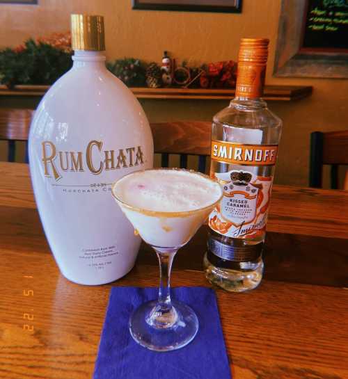 A cocktail glass with a creamy drink, surrounded by bottles of RumChata and Smirnoff Kissed Caramel on a wooden table.