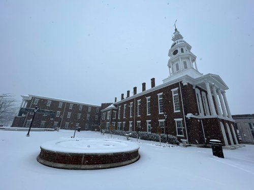 A snowy scene featuring a historic building with a clock tower and a circular fountain in the foreground.