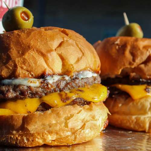 Two juicy cheeseburgers with melted cheese, topped with olives, on a wooden surface.
