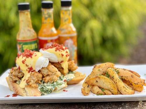 A hearty sandwich with fried chicken, poached eggs, and hollandaise sauce, served with seasoned potato wedges and hot sauces.
