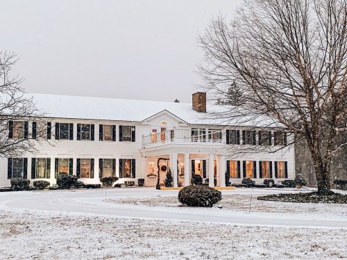 A large white house with black shutters, surrounded by snow, featuring warm lights and a festive wreath on the door.
