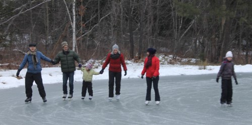 A group of six people ice skating on a frozen pond, surrounded by trees in winter attire.