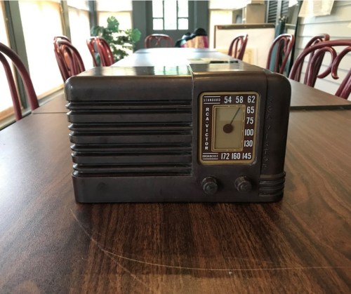 A vintage radio with a dial and knobs, placed on a wooden table in a room with chairs in the background.