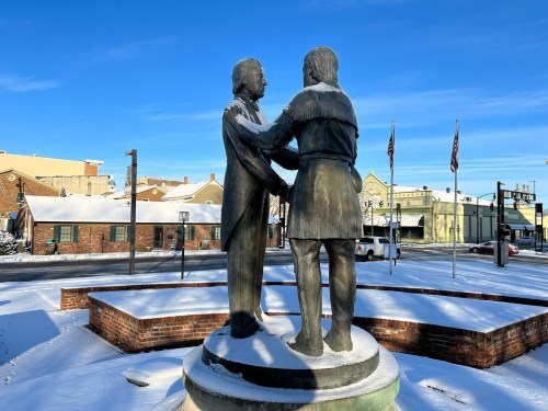 Statue of two figures in winter, covered in snow, with a clear blue sky and buildings in the background.
