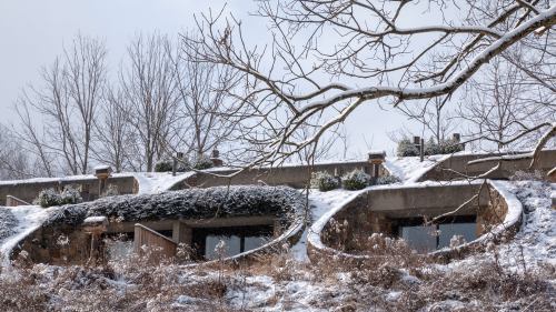 A snow-covered hillside with modern, curved buildings partially embedded in the landscape, surrounded by bare trees.