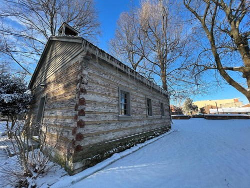 A rustic log cabin with icicles, surrounded by snow and bare trees under a clear blue sky.