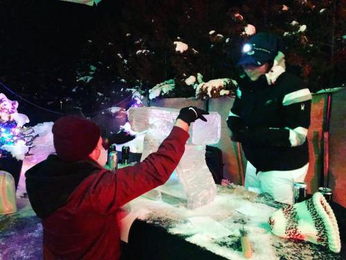 Two people work on an ice sculpture outdoors, surrounded by snow and festive lights.