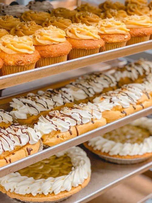 A display of cupcakes with frosting, cream-filled pastries, and pies on metal shelves in a bakery.