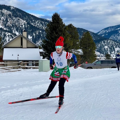 A skier in a festive outfit races on snow-covered trails with mountains in the background.