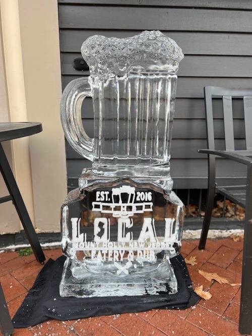 An ice sculpture of a beer mug with "LOCAL" and "EST. 2016" carved into it, placed outside on a black mat.