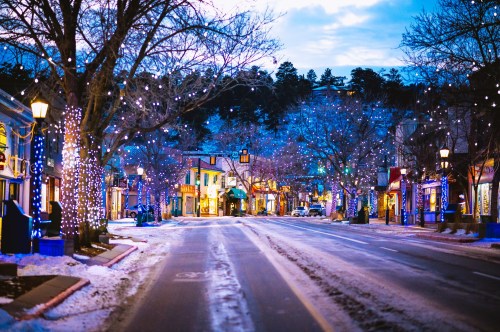 A snowy street lined with trees adorned with colorful lights, creating a festive atmosphere at dusk.