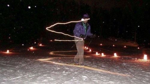 A skier glides through a snowy landscape at night, leaving a trail of light amidst glowing lanterns.