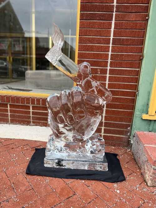 A clear ice sculpture of a raised fist holding a paintbrush, displayed on a black cloth outside a brick building.