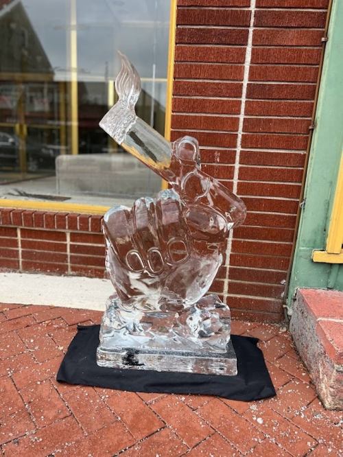 A clear ice sculpture of a raised fist holding a paintbrush, displayed on a black cloth outside a brick building.