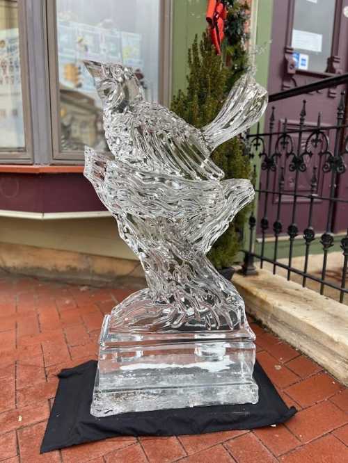 A detailed ice sculpture of a bird perched on a branch, displayed on a black cloth outside a building.