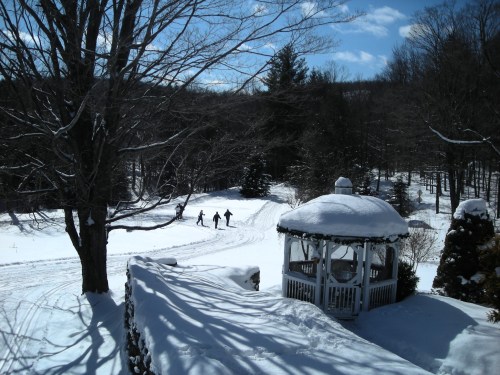 A snowy landscape with a gazebo and four people walking in the distance under a bright blue sky.