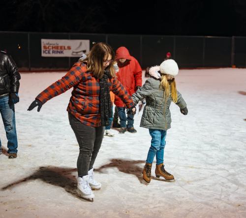 A woman and a girl skate on ice at night, surrounded by others in winter clothing. Snowy rink atmosphere.