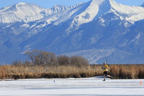 A person ice skating on a frozen lake with snow-capped mountains in the background and tall grasses nearby.