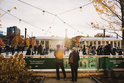 People skate on an outdoor rink surrounded by autumn trees and festive lights, with a sunset in the background.