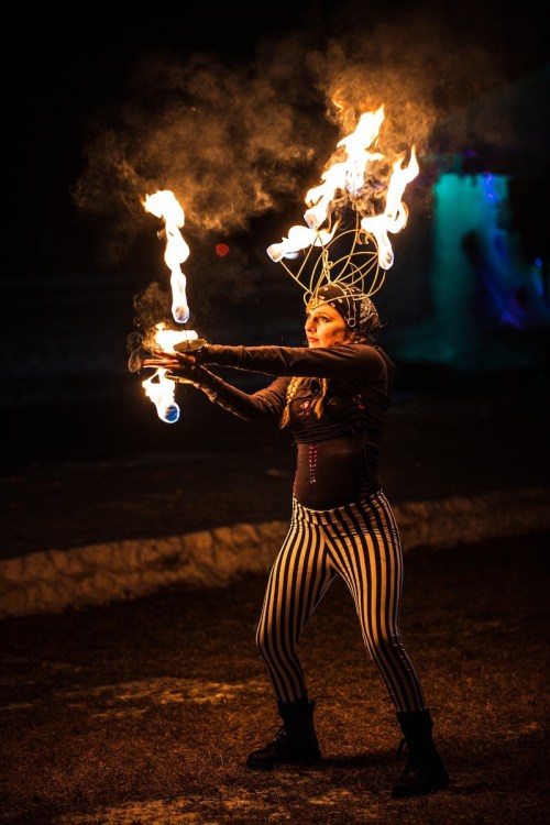A performer in striped pants juggles flaming torches at night, with smoke and fire illuminating the scene.