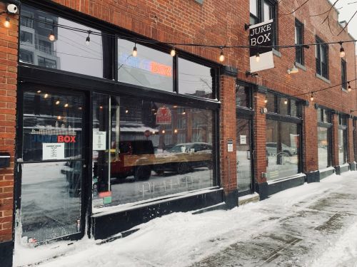 A brick building with large windows, featuring a sign that reads "Juke Box," surrounded by snow on the ground.