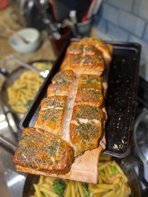A wooden plank with grilled salmon fillets, garnished with herbs, on a kitchen counter with pasta in the background.