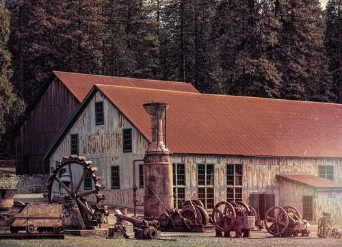 Rustic building with a red roof, surrounded by vintage machinery and trees in the background.