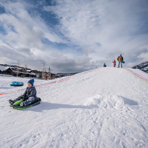 A child sleds down a snowy hill while others enjoy the winter landscape in the background.