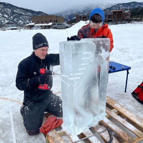 Two people sculpting a large block of ice outdoors, with mountains in the background and tools on a table nearby.