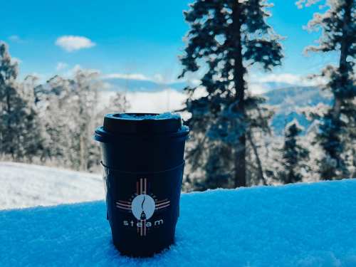 A coffee cup sits on snow, surrounded by snowy trees and a clear blue sky in a winter landscape.