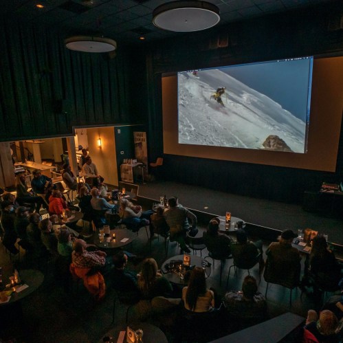 A crowd watches a skiing film on a large screen in a cozy theater setting with tables and drinks.