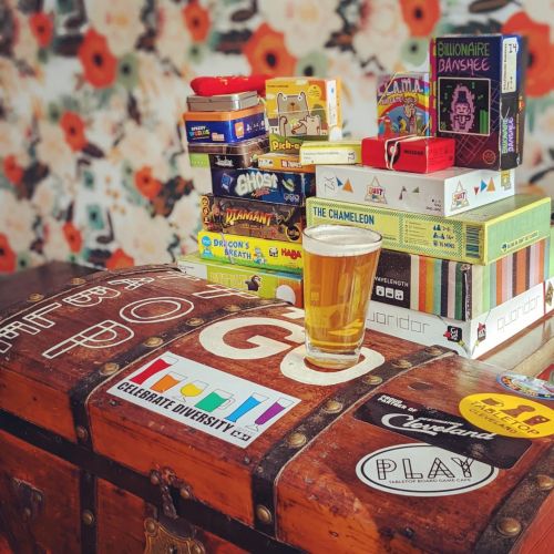 A wooden trunk filled with board games and a glass of beer, set against a floral wallpaper background.