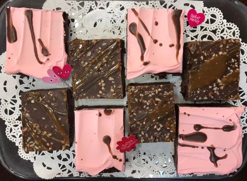 A tray of chocolate and pink frosted brownies, decorated with heart-shaped picks and placed on a lace doily.