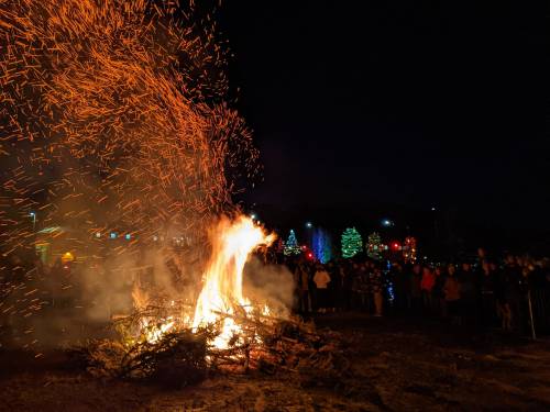 A large bonfire with sparks flying, surrounded by a crowd enjoying a festive night with decorated trees in the background.