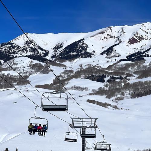 A ski lift carries passengers over a snowy landscape with mountains in the background under a clear blue sky.