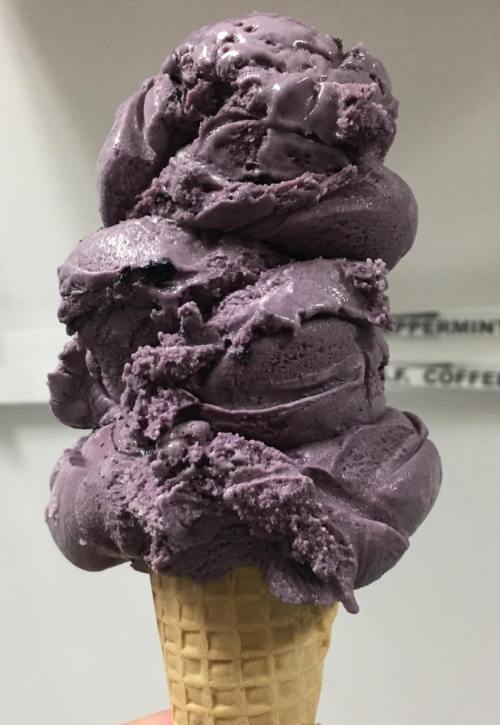 A tall cone of purple ice cream with a smooth, creamy texture, topped with swirls of the same color.