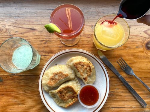 A plate of golden-brown dumplings with dipping sauce, surrounded by colorful cocktails on a wooden table.