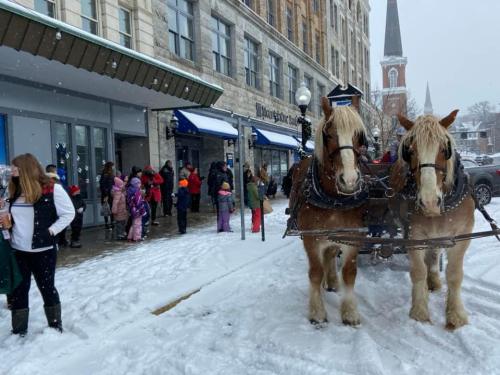 Two horses in harness stand in the snow outside a building, with a line of people and children in winter clothing nearby.