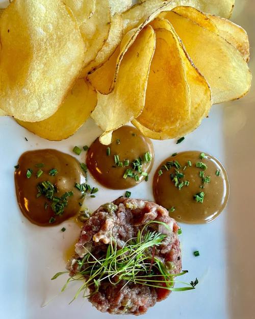 A plate featuring crispy potato chips, beef tartare topped with microgreens, and three sauces garnished with herbs.