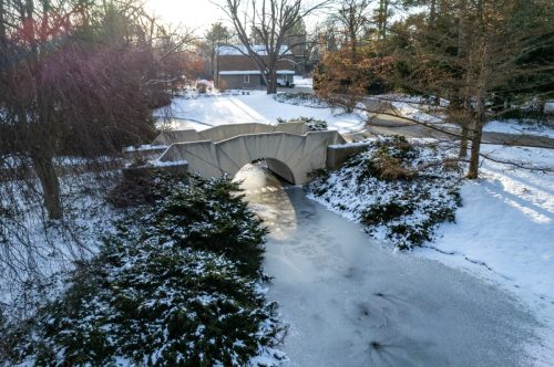 A snowy landscape featuring a small bridge over a frozen stream, surrounded by trees and a residential area.