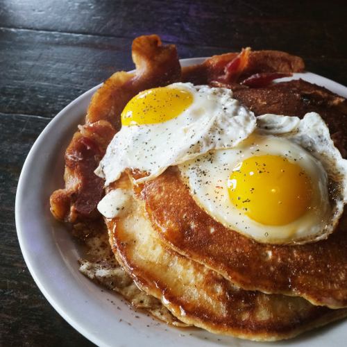A plate of pancakes topped with two sunny-side-up eggs and crispy bacon on the side.