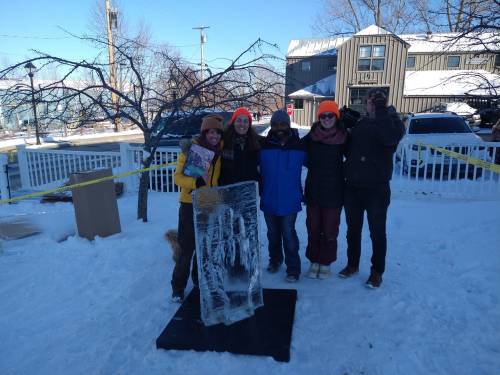 Five people stand in the snow near an ice sculpture, wearing winter clothing and bright orange hats, smiling at the camera.