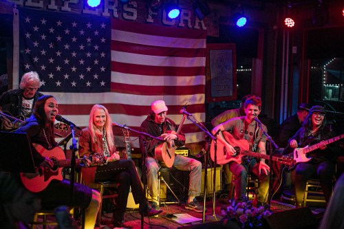 A lively music performance with six musicians playing guitars in front of an American flag backdrop.