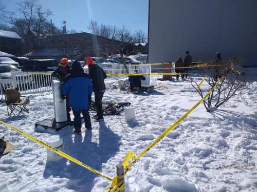 Workers in winter gear set up equipment in a snowy area, surrounded by caution tape and a white fence.
