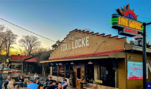 A lively outdoor scene at Fox & Locke, featuring people dining and enjoying live music under a colorful sunset.