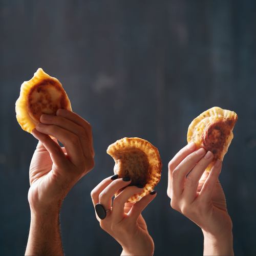 Three hands hold up golden-brown empanadas against a dark background, showcasing their crispy edges.