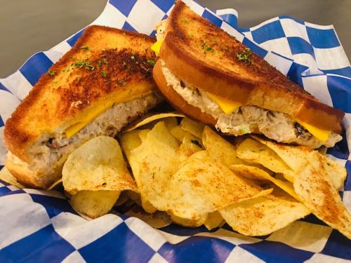 Grilled cheese sandwich with tuna salad, served with crispy potato chips on a blue checkered paper.